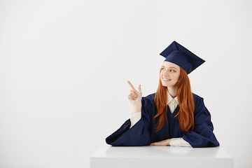 young-female-university-graduate-in-academic-cap-sitting-at-table-smiling-pointing-left-future-lawyer-or-engineer-showing-an-idea_176420-14245